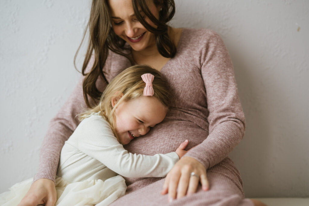 A two-year-old girl hugs her mom's pregnant belly during maternity portraits in a Denver photo studio