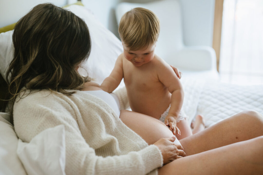 A little boy in a diaper touches his mom's pregnant belly during maternity portraits in their Denver home.
