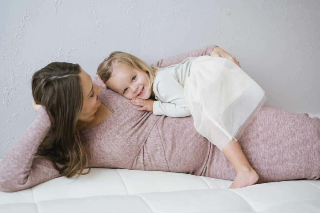 A pregnant mom lies with her little daughter during in studio maternity portraits in Denver.