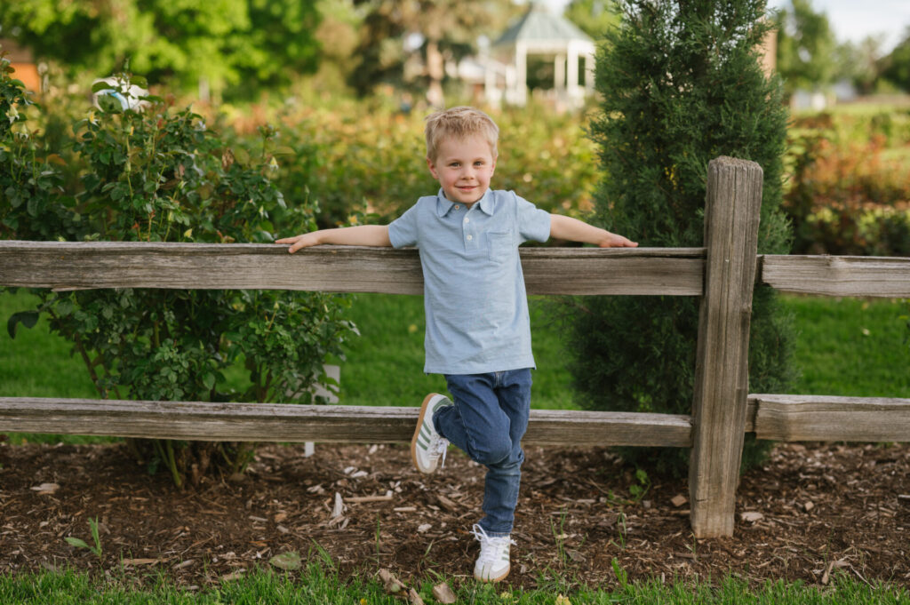 A little boy leans against a fence with the rose garden in the back.